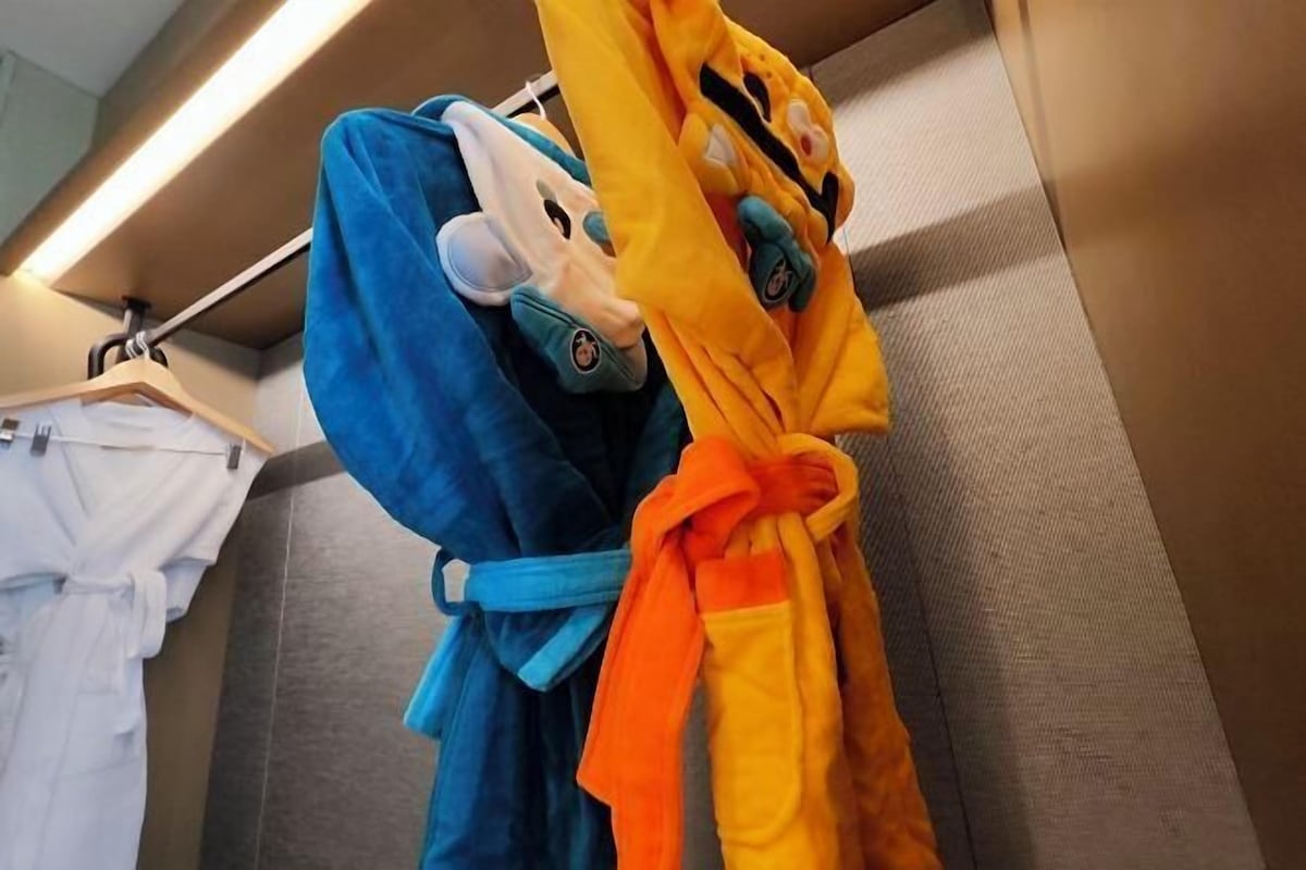 A hotel room with a grey wall and a wooden hanger holding a blue robe and an orange robe.