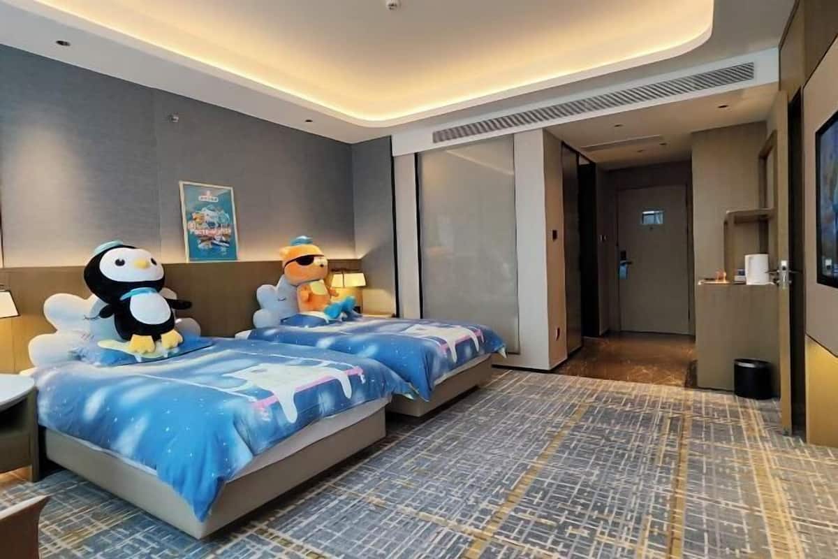 A hotel room with two beds, each with a penguin plush toy, a poster on the wall, and a TV on the right.