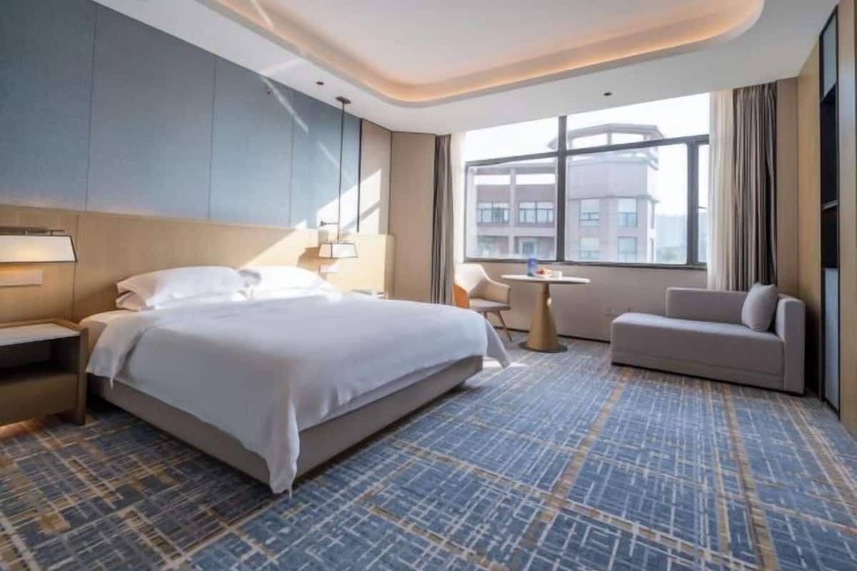 A modern hotel room with a large bed, a desk, a sofa, and a view of the city through large windows.