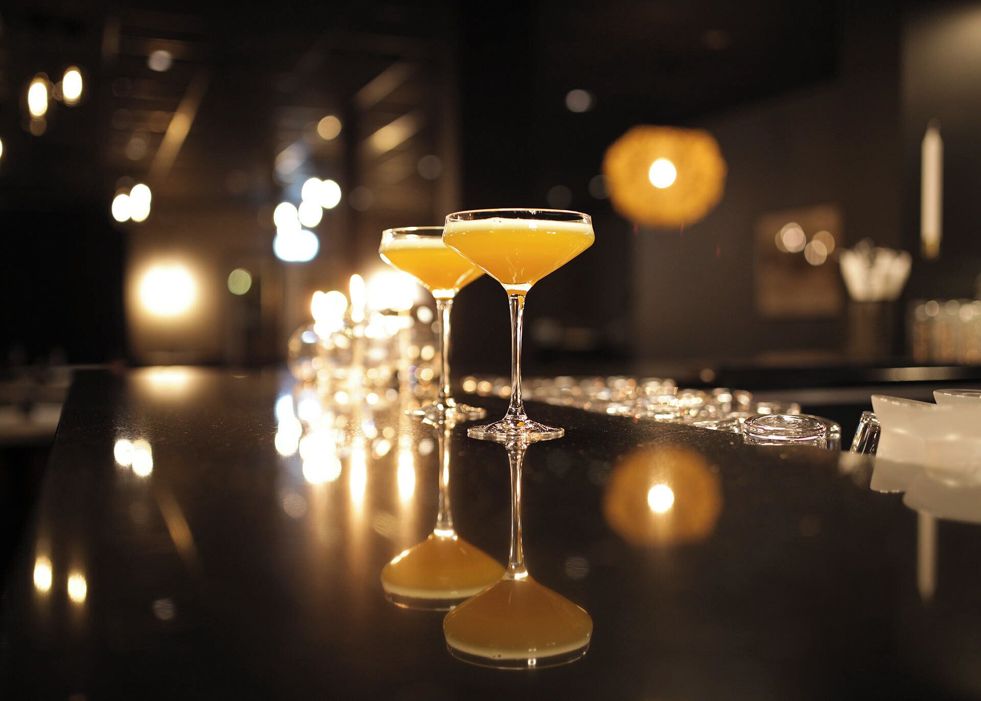 Dos vasos de martini con una bebida amarillenta sobre una barra reflectante.