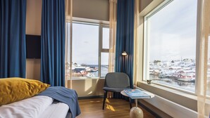 A hotel room with a bed, a chair, a table, and a view of a snowy village through large windows. - The Tide - Hotel (Moskenes)