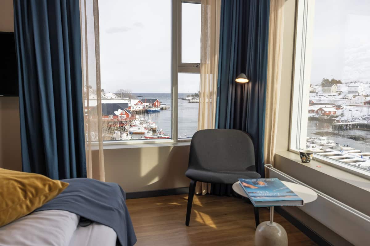 A hotel room with a bed, a chair, a small table with a book, and a window offering a view of a harbor with boats and buildings.