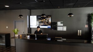 A reception desk with a person seated behind it, a large screen displaying 'THE TIDE' advertisement, and a potted plant on the left. - The Tide - Hotel (Moskenes)
