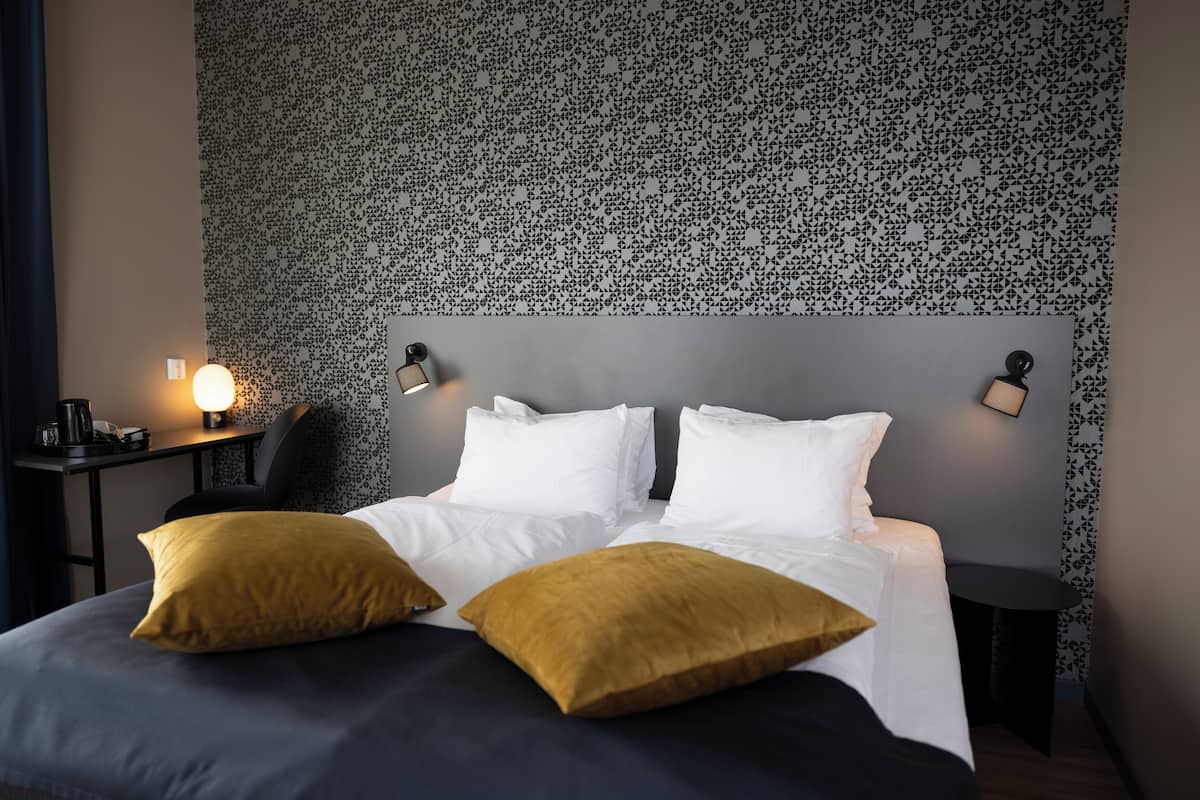 A neatly made bed with white and mustard pillows, a grey headboard, and patterned wallpaper.