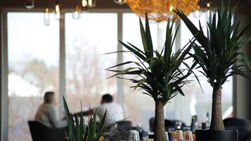 Un interior de restaurante con ventanas amplias, plantas en macetas y luces colgantes.