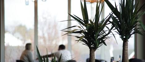 A restaurant interior with large windows, potted plants, and hanging lights.