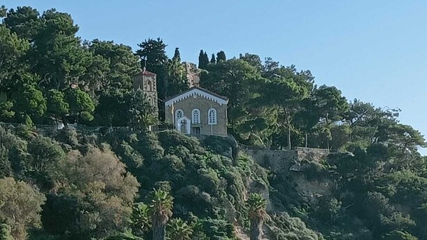 A coastal building with a bell tower, surrounded by dense greenery and rocky cliffs.