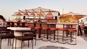 An outdoor bar area with multiple seating options, including round tables and bar stools, under large umbrellas.