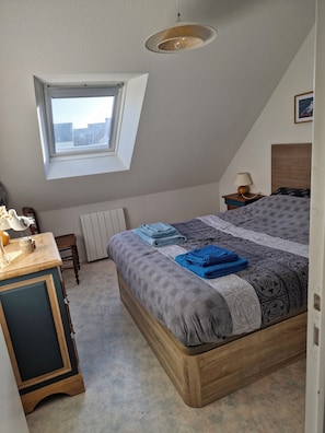 A bedroom with a sloped ceiling, a window, a bed with patterned bedding, a bedside table with a lamp, and a radiator.