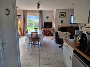 Uma sala de jantar com mesa e cadeiras, uma cozinha com eletrodomésticos e vista através de uma porta de vidro.