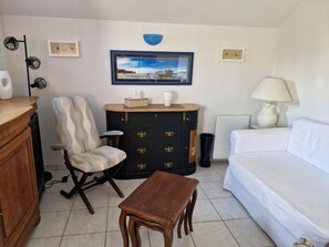 Uma sala de estar com sofá branco, mesa de madeira, aparador preto com puxadores decorativos, um quadro emoldurado na parede e um abajur.