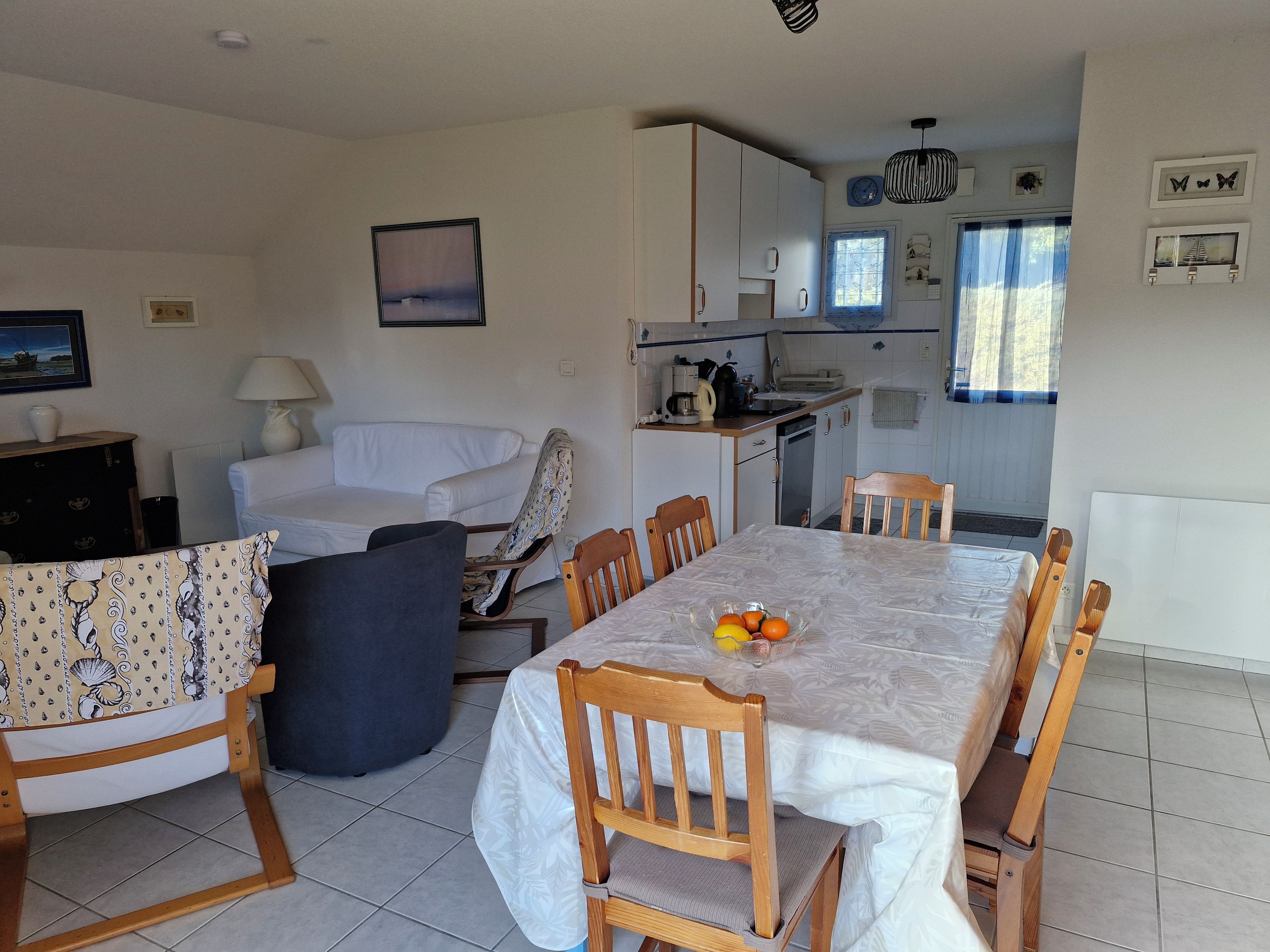 A dining area with a table set for six, surrounded by wooden chairs. A kitchen is visible in the background with a window and curtains.