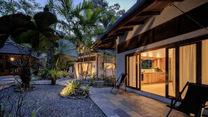 Ein modernes Haus mit Terrasse, umgeben von tropischen Pflanzen und einem Kiesweg.