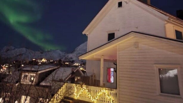 Une maison avec un toit en pente et une cheminée bien visible, entourée de montagnes enneigées et offrant une vue sur les aurores boréales.