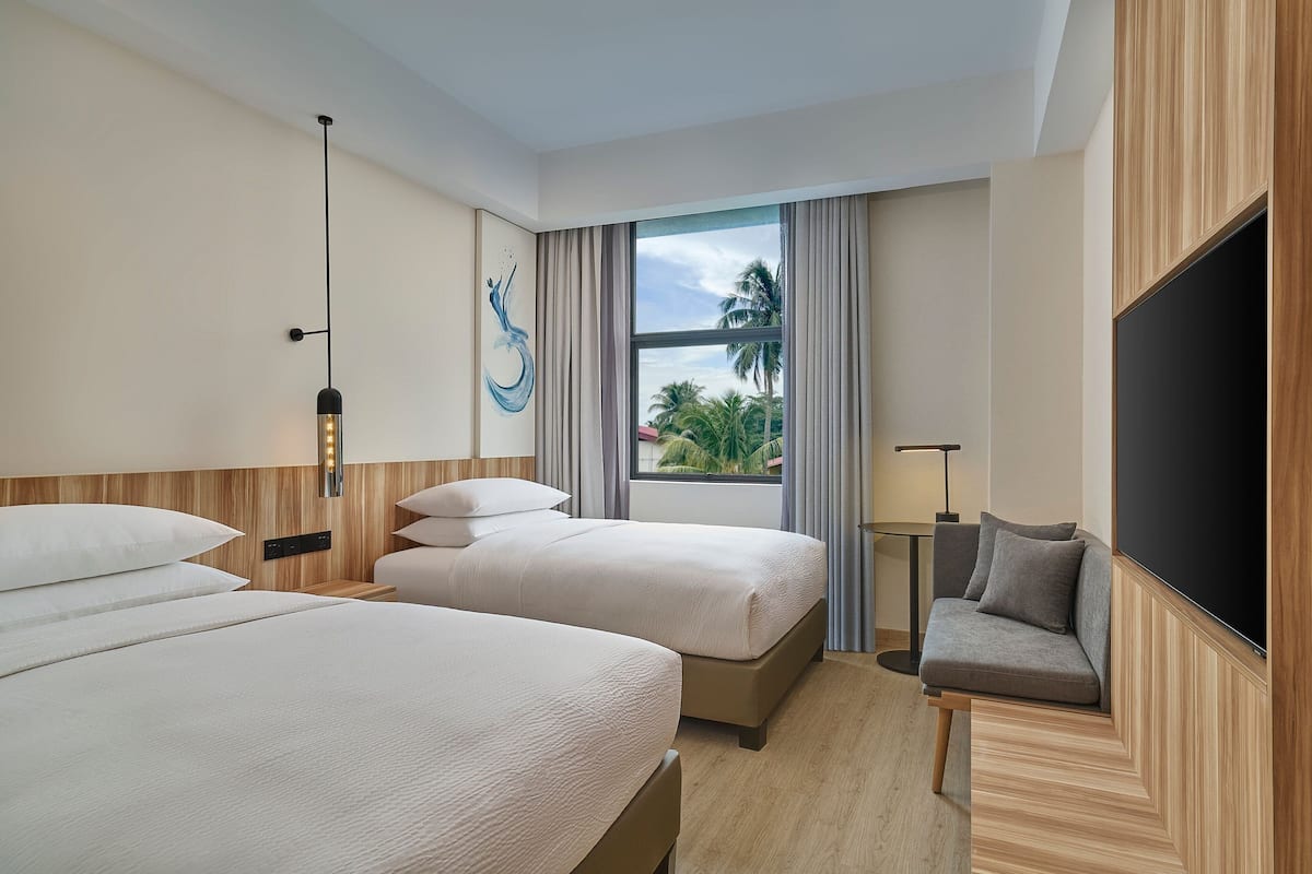 A hotel room with two beds, a sofa, a TV, and a window with a view of palm trees.