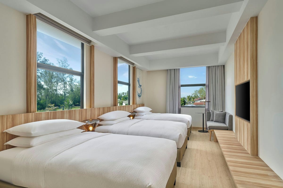 A hotel room with three beds, a TV, and large windows offering a view of trees and a building.
