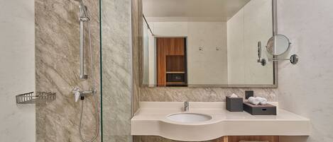 A modern bathroom with a glass-enclosed shower, a marble-tiled wall, a sink with a mirror, and a wooden shelf with towels.