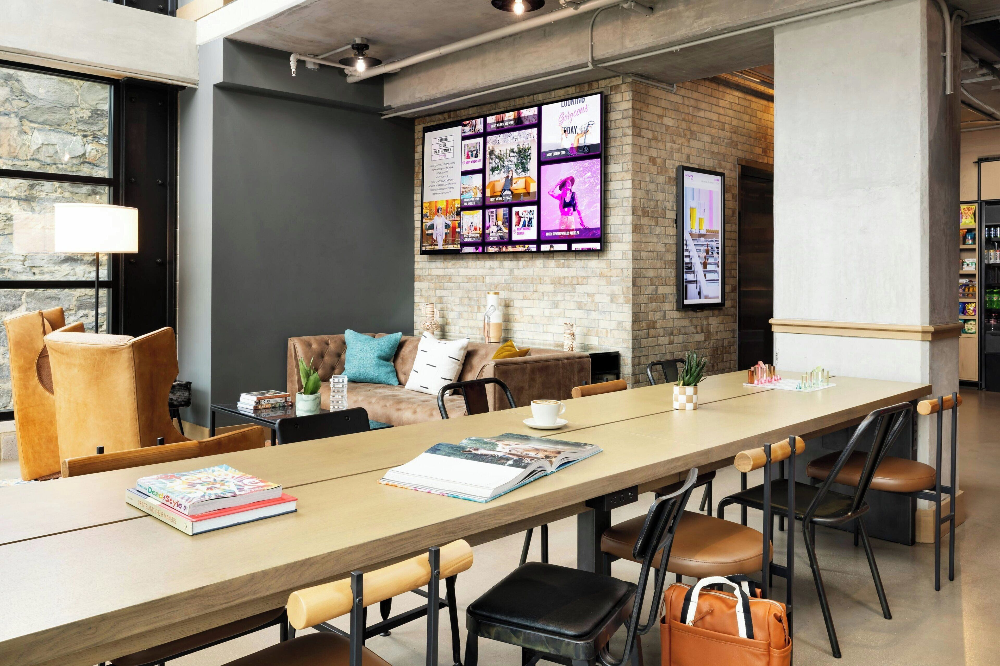 A modern meeting room with a long table, comfortable seating, a brick wall with a digital display, and a stone wall with a window.