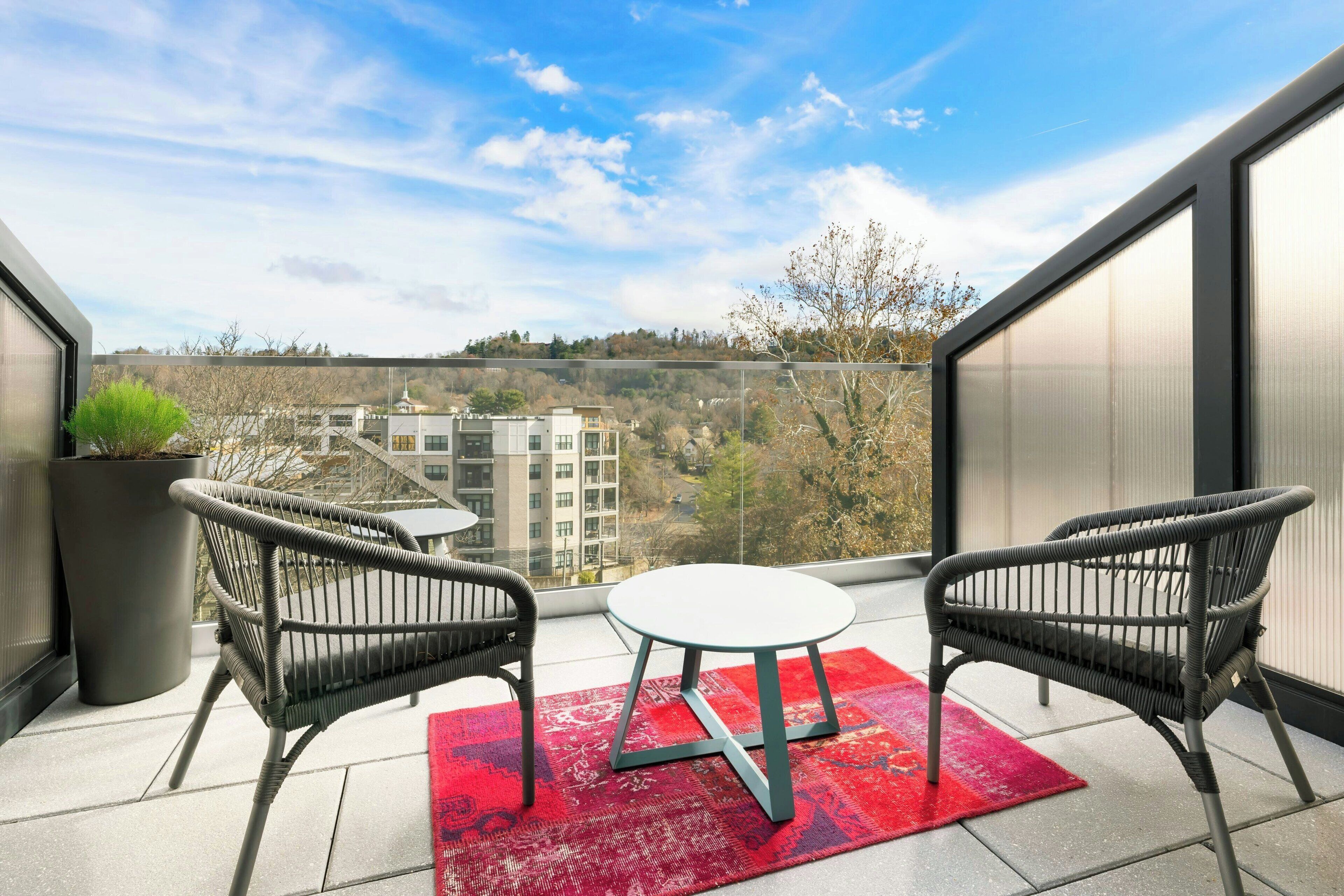 A balcony with two wicker chairs and a small round table, a red rug, and a view of buildings and trees.