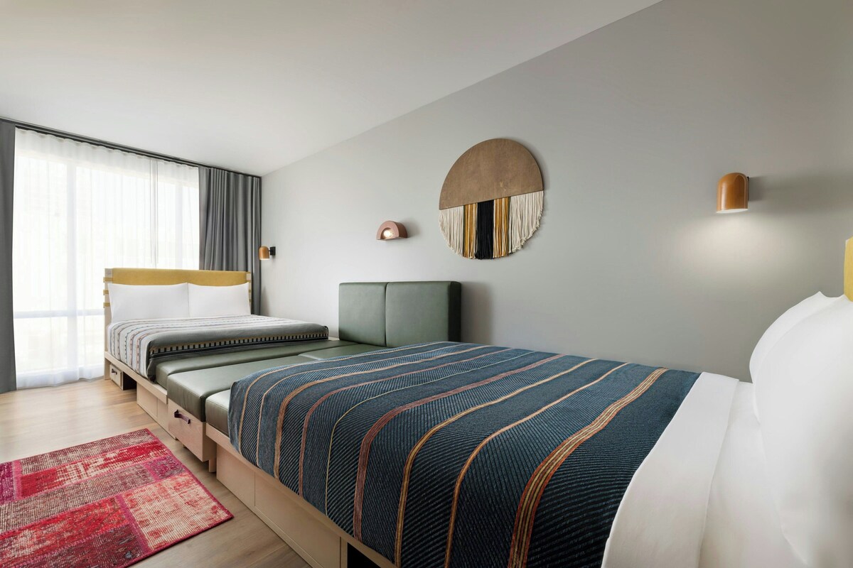 A modern hotel room with a bed, a bedside table, a lamp, a round wall decoration, and a window with curtains.