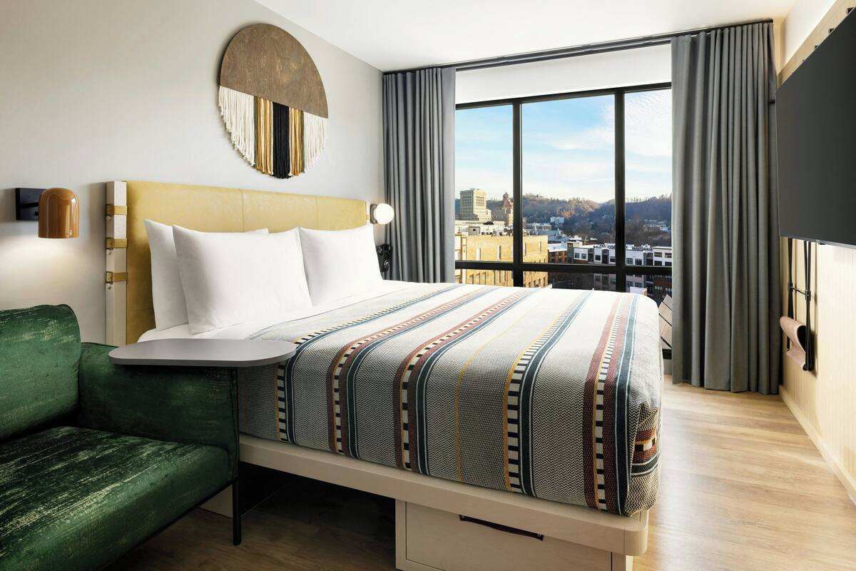 A modern hotel room with a large bed, a green sofa, a city view, and a large window with curtains.