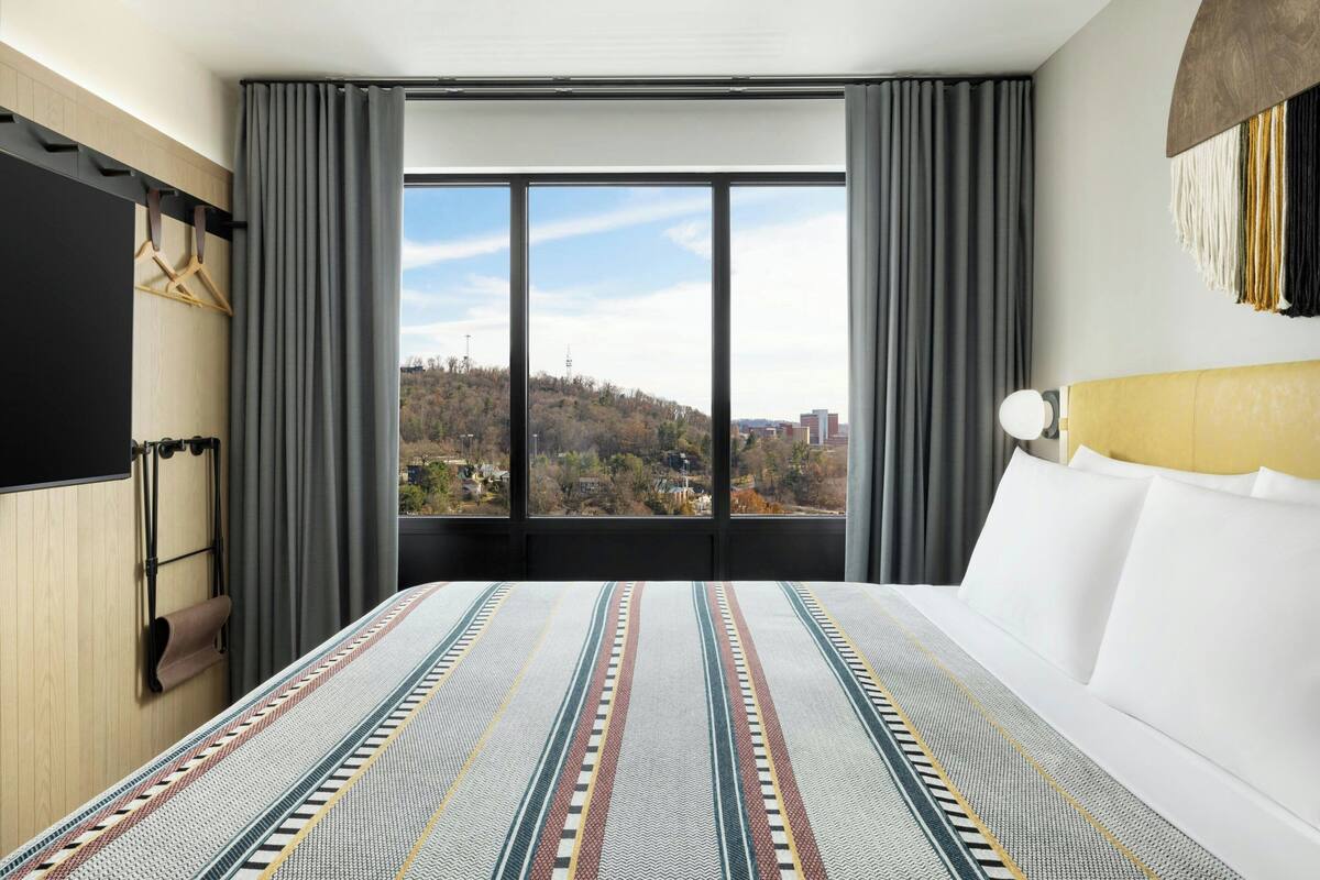 A hotel room with a large bed, a TV mounted on the wall, and a window with a view of a cityscape.