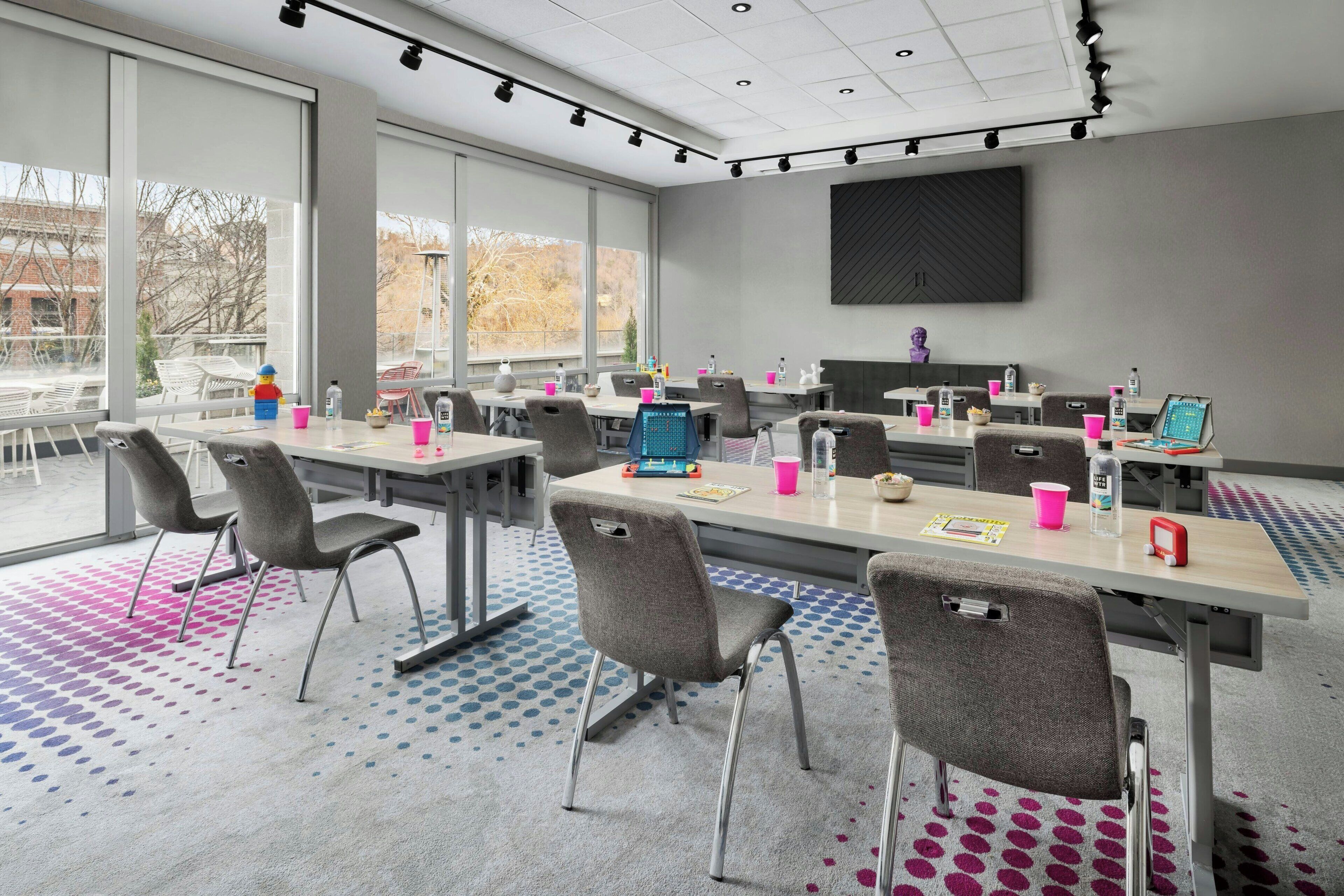 A conference room with large windows, multiple tables, and chairs, equipped with laptops and various office supplies.