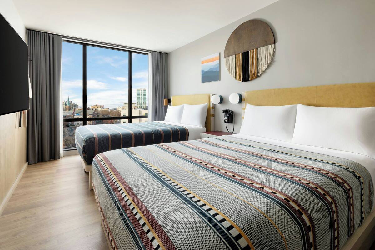 A hotel room with two beds, a large window with a city view, a round wall decoration, and a flat-screen TV.