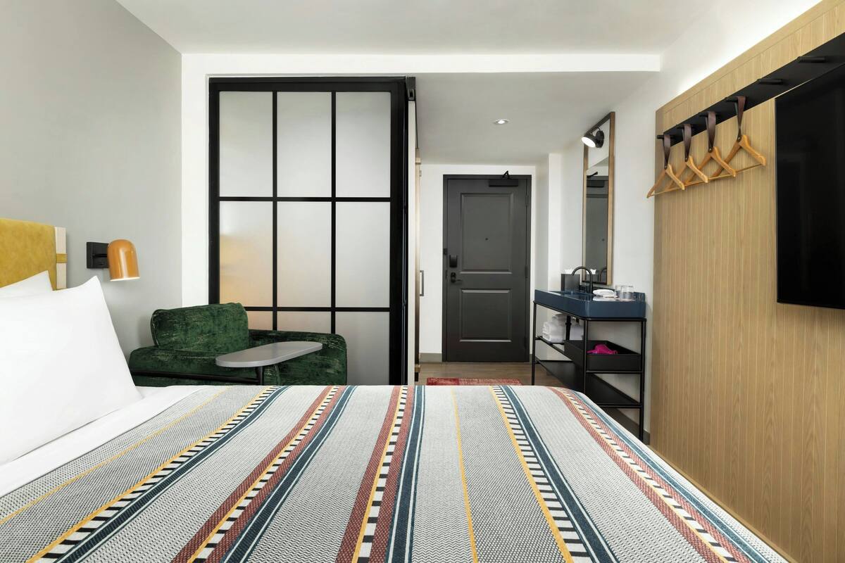 A modern hotel room with a large bed, a green sofa, a small table, a wooden wardrobe, a mirror, and a television.