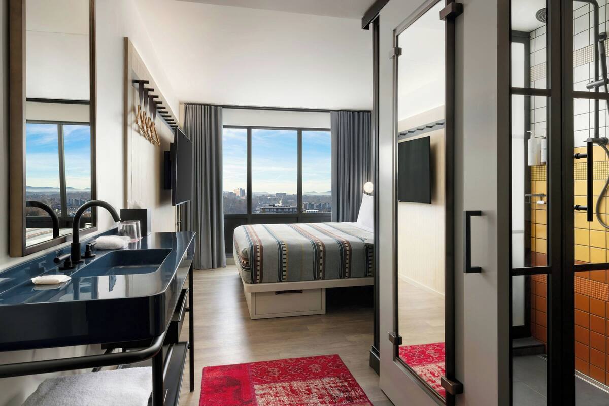A modern hotel room with a large bed, a bathroom with a shower, a sink, and a city view.