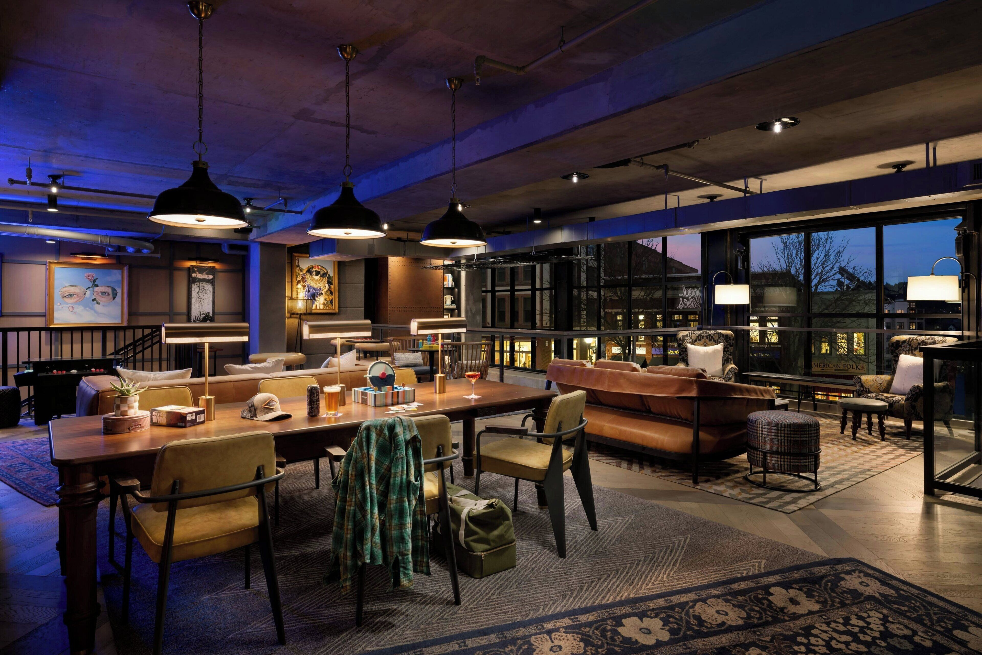 Un hall d’hôtel moderne et bien éclairé, doté d’une grande table à manger, de sièges confortables et de larges fenêtres offrant une vue sur le paysage urbain.