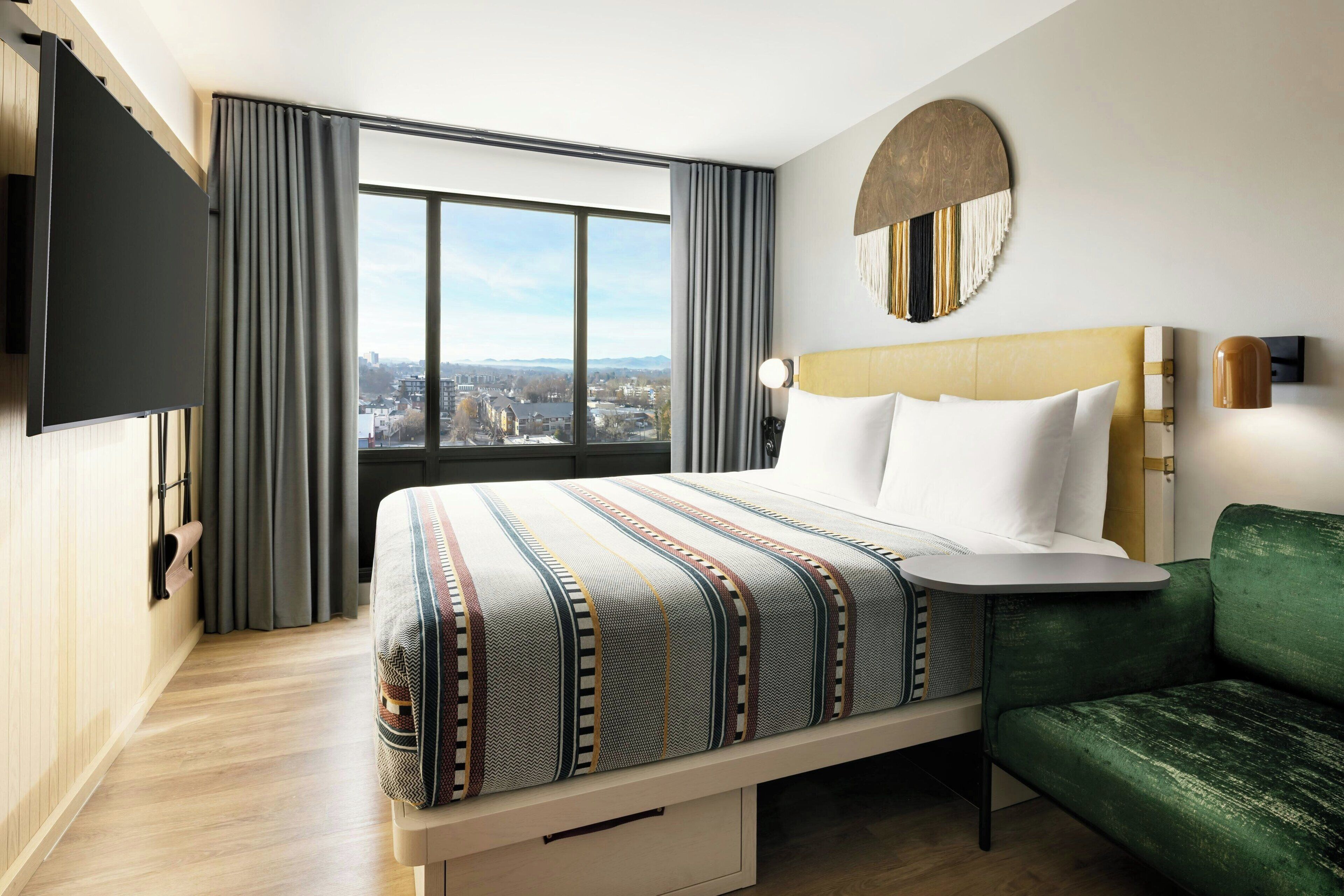 A hotel room with a large bed, a green sofa, a TV mounted on the wall, and a city view through the window.