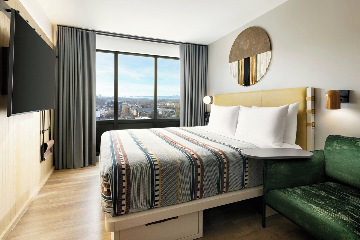 A hotel room with a large bed, a green sofa, a TV mounted on the wall, and a city view through the window.
