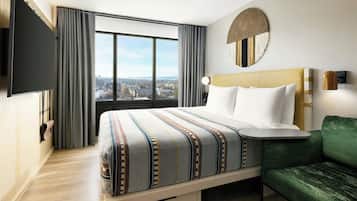 Une chambre d’hôtel avec un grand lit, un canapé vert, un téléviseur fixé au mur et une vue sur la ville par la fenêtre.