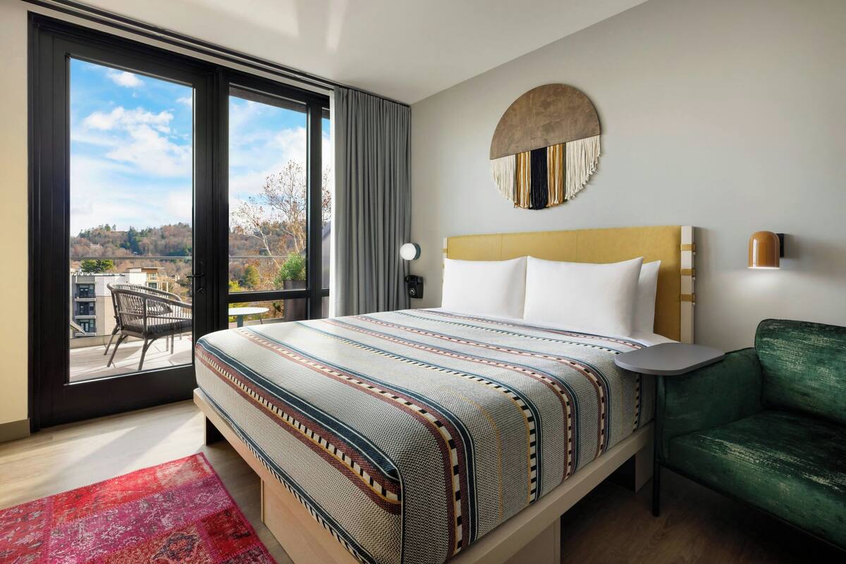 A hotel room with a large bed, a green sofa, a round wall decoration, and a view of a balcony with a chair.