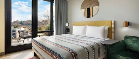A hotel room with a large bed, a green sofa, a round wall decoration, and a view of a balcony with a chair.