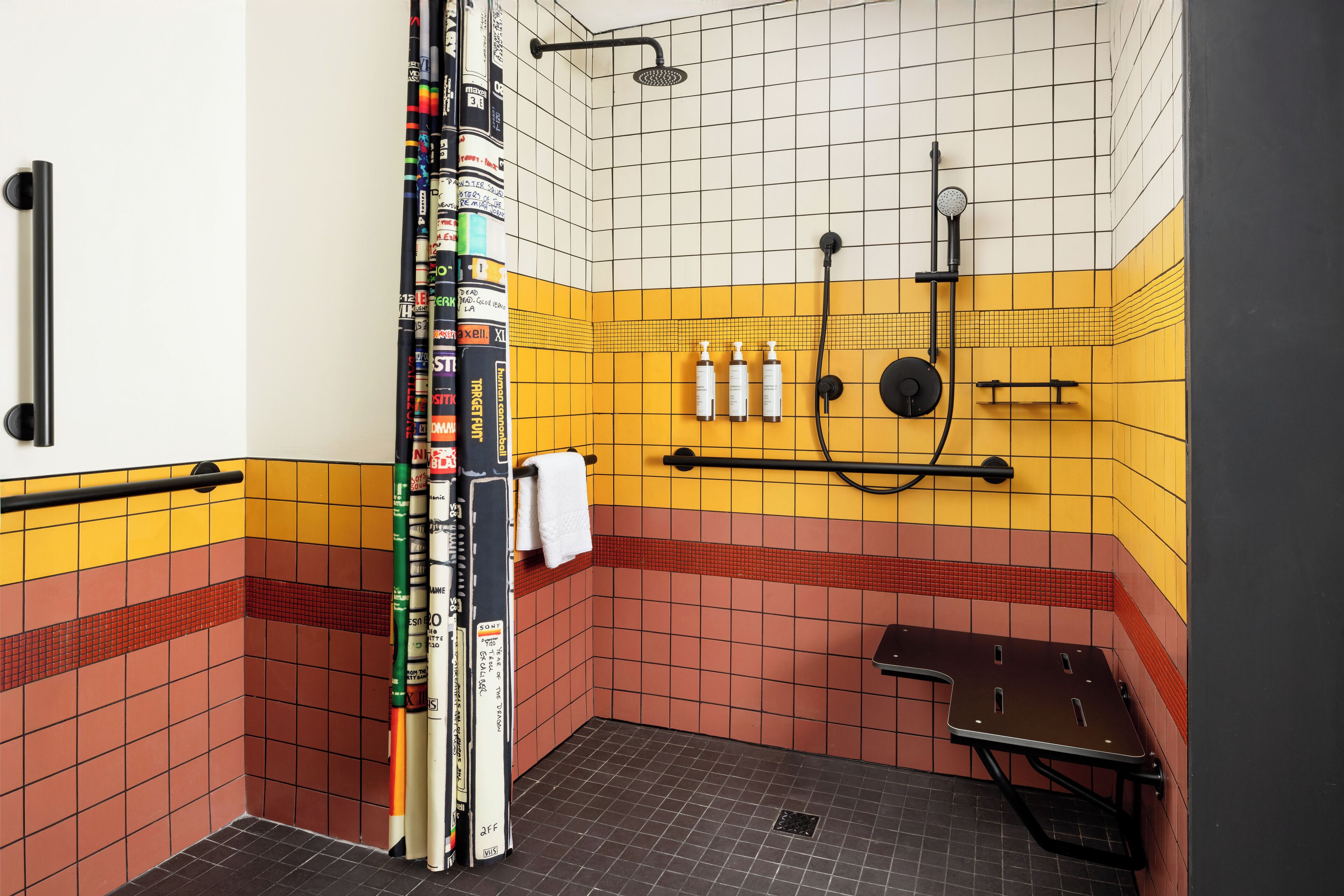 A small bathroom with yellow and red tiles, a shower, a towel rack, and a shelf with toiletries.
