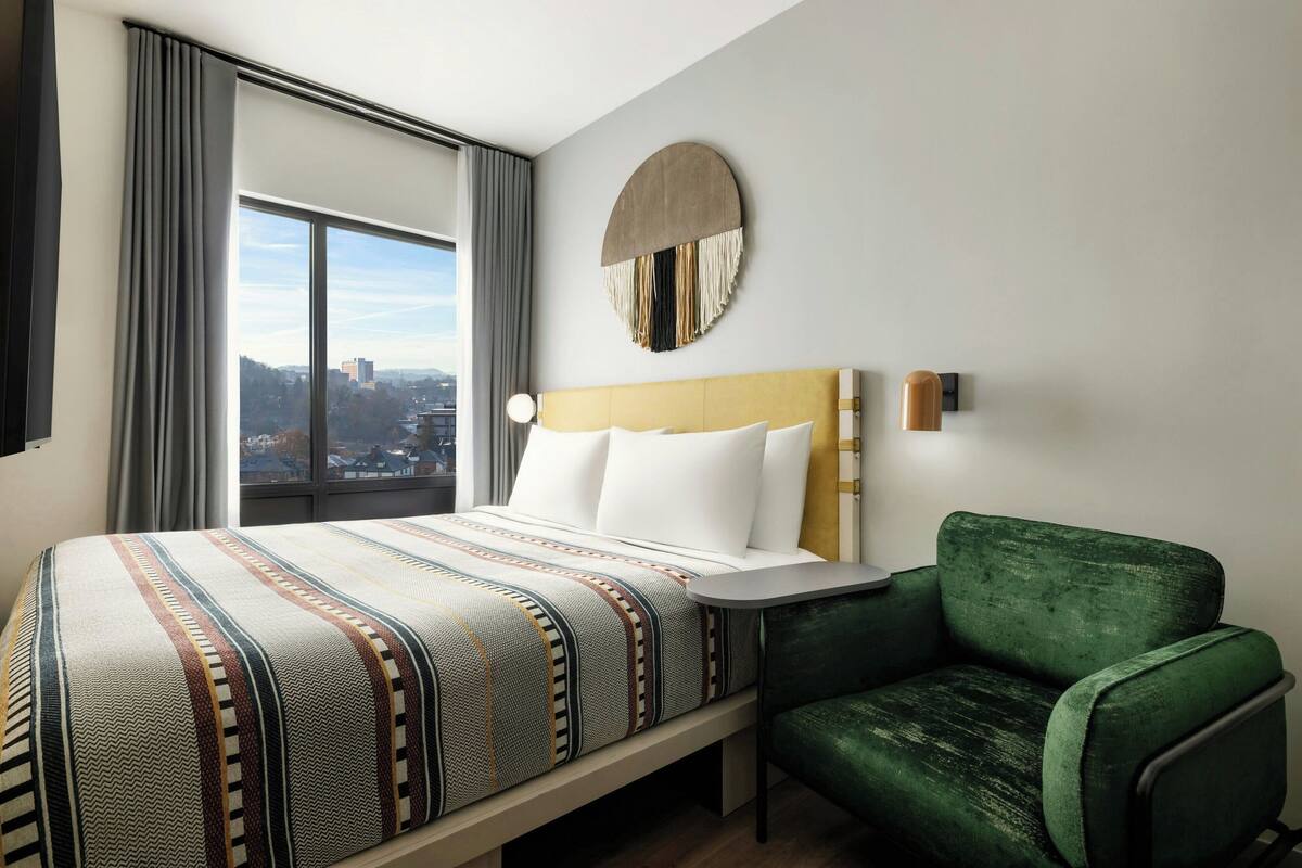 A hotel room with a large bed, a green armchair, a round wall decoration, and a window with a city view.