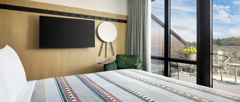 Ein Hotelzimmer mit einem großen Bett, einem an der Wand montierten Fernseher, einem runden Spiegel, einem grünen Sessel und einem Balkon mit Blick ins Freie.