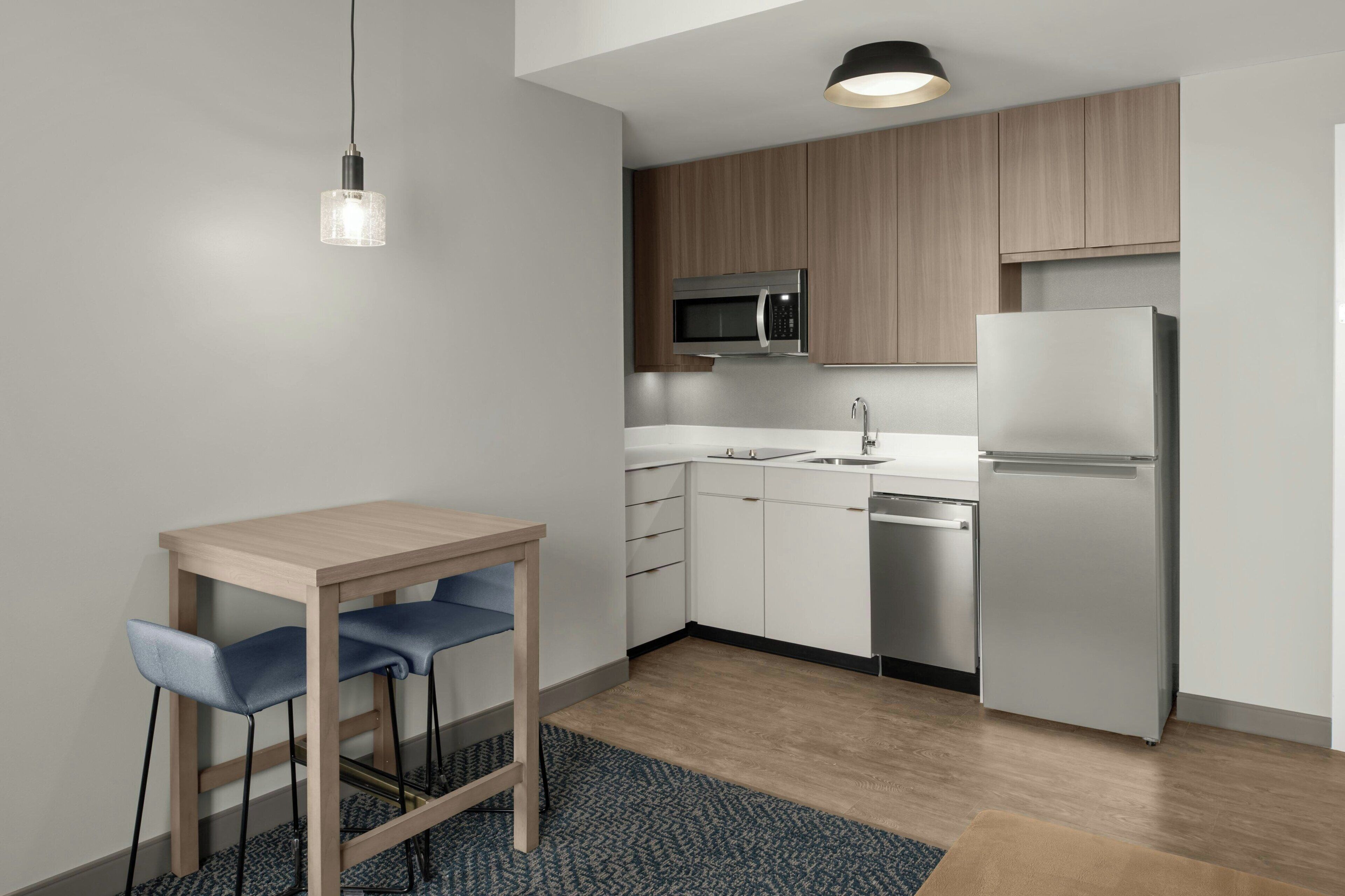 A modern kitchen with white cabinets, a stainless steel refrigerator, a microwave, and a sink. There is a wooden table with two chairs and a hanging light fixture.