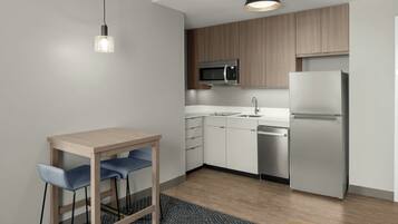 A modern kitchen with white cabinets, a stainless steel refrigerator, a microwave, and a sink. There is a wooden table with two chairs and a hanging light fixture.