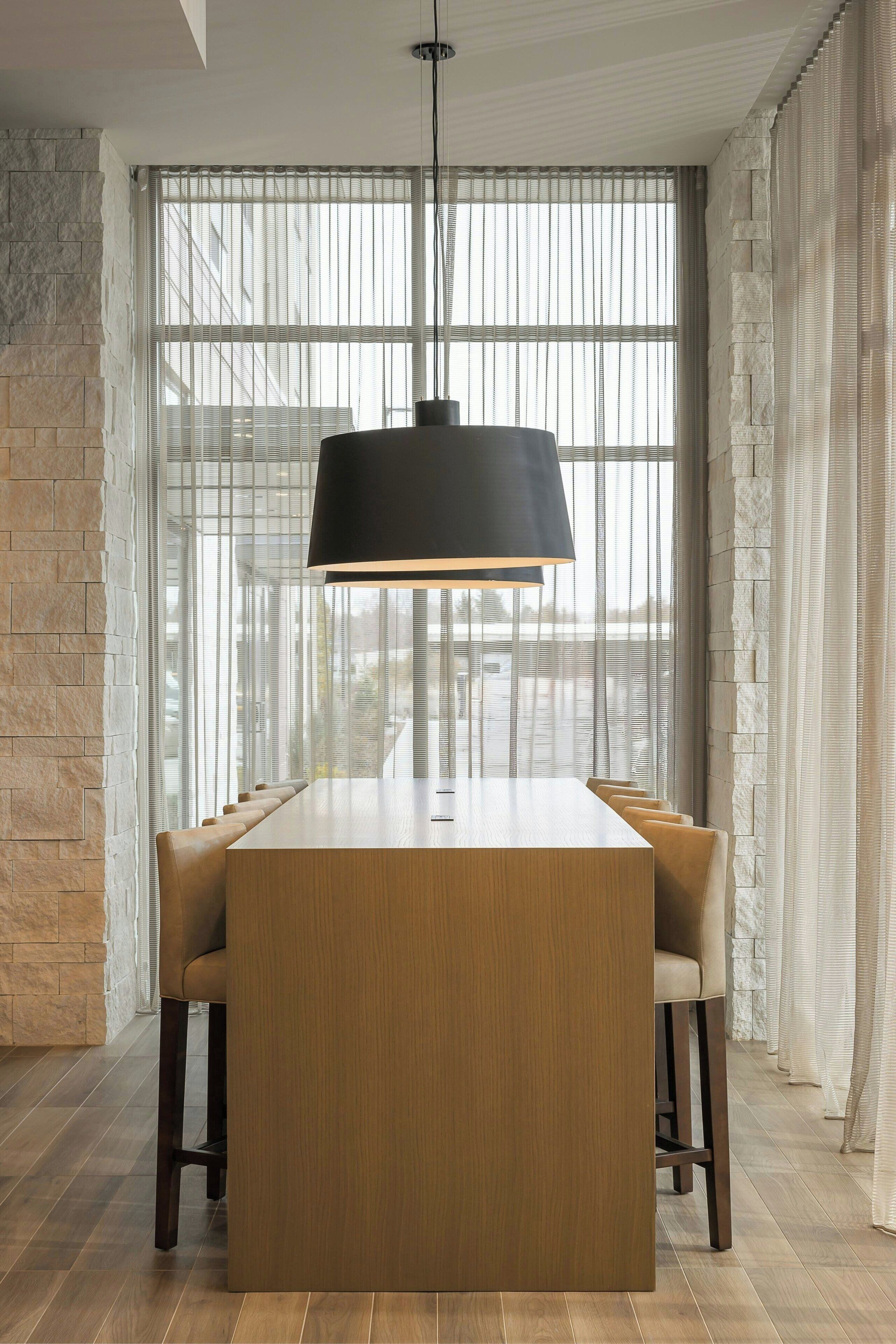A modern dining area with a long wooden table and cushioned chairs, a black pendant light, and a large window with sheer curtains.