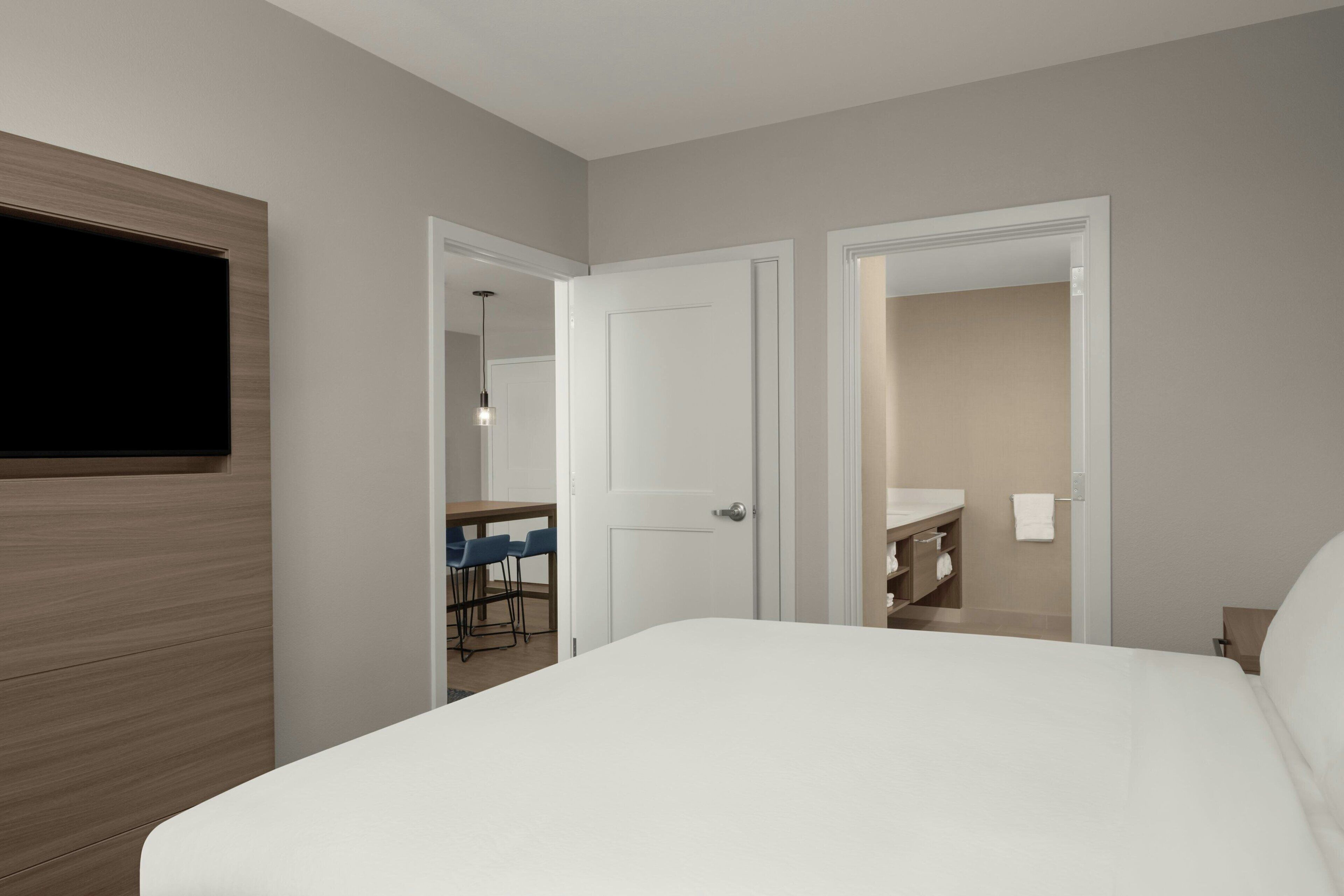Una habitación de hotel moderna con una cama grande, un televisor de pantalla plana montado en la pared y un área de comedor separada visible a través de una puerta abierta.