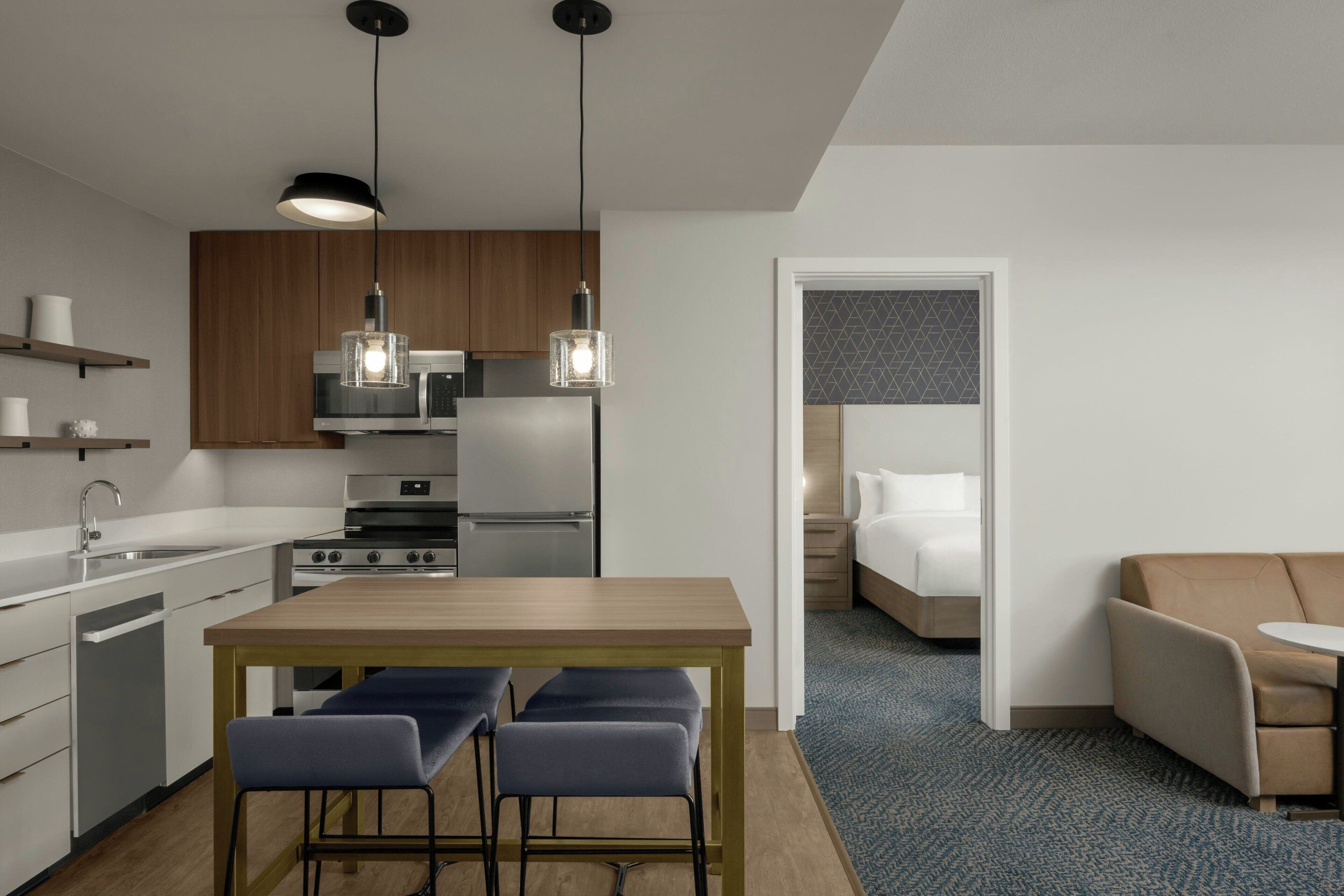A modern hotel room with a kitchenette, a dining area with a wooden table and bar stools, a bed with a white headboard, and a blue carpet.