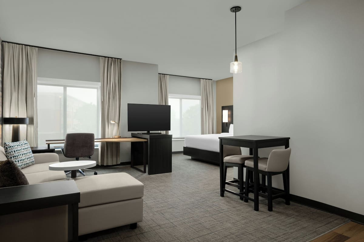 Una habitación de hotel moderna con sofá, una mesita redonda pequeña, televisor, cama y escritorio con dos sillas.