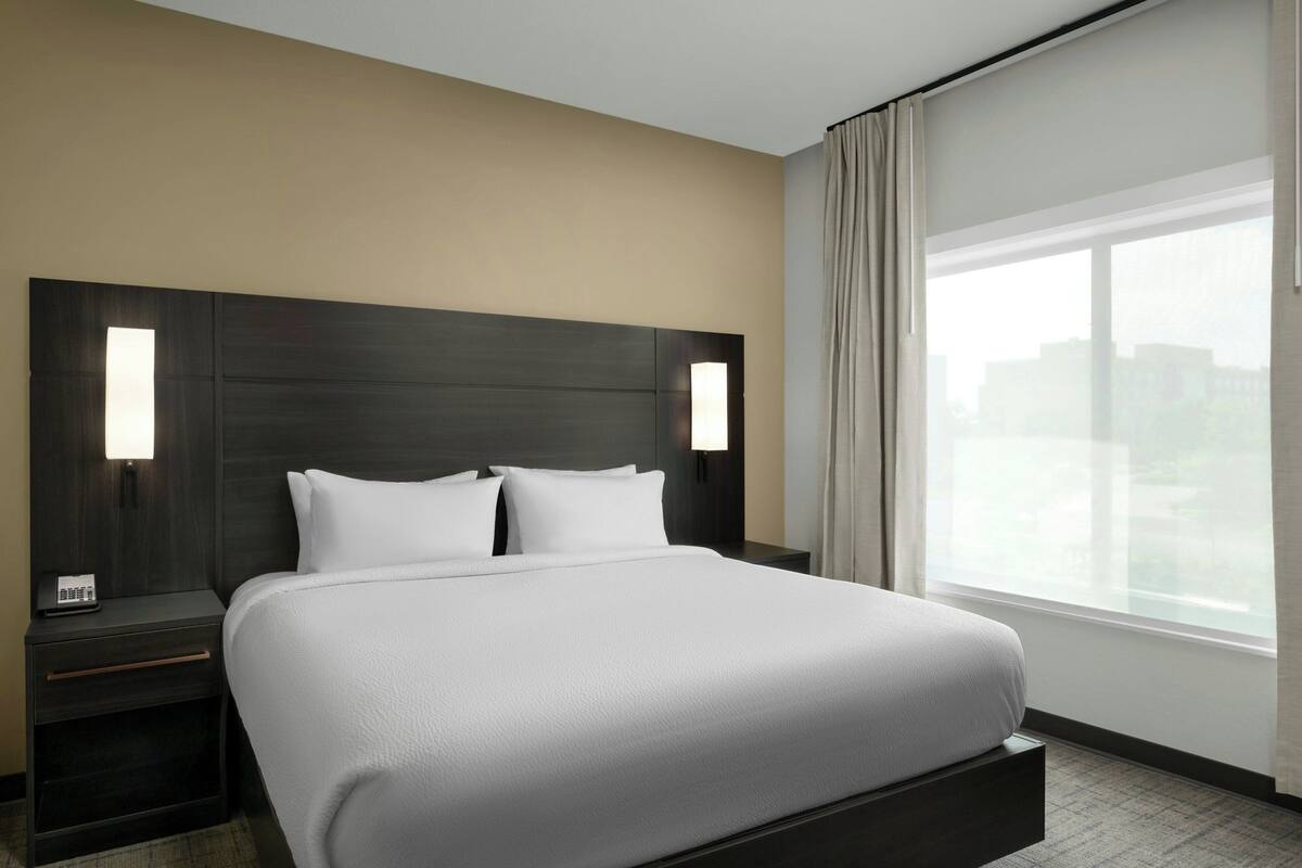 Una habitación de hotel moderna con una cama grande, dos mesitas de noche y un ventanal con cortinas.