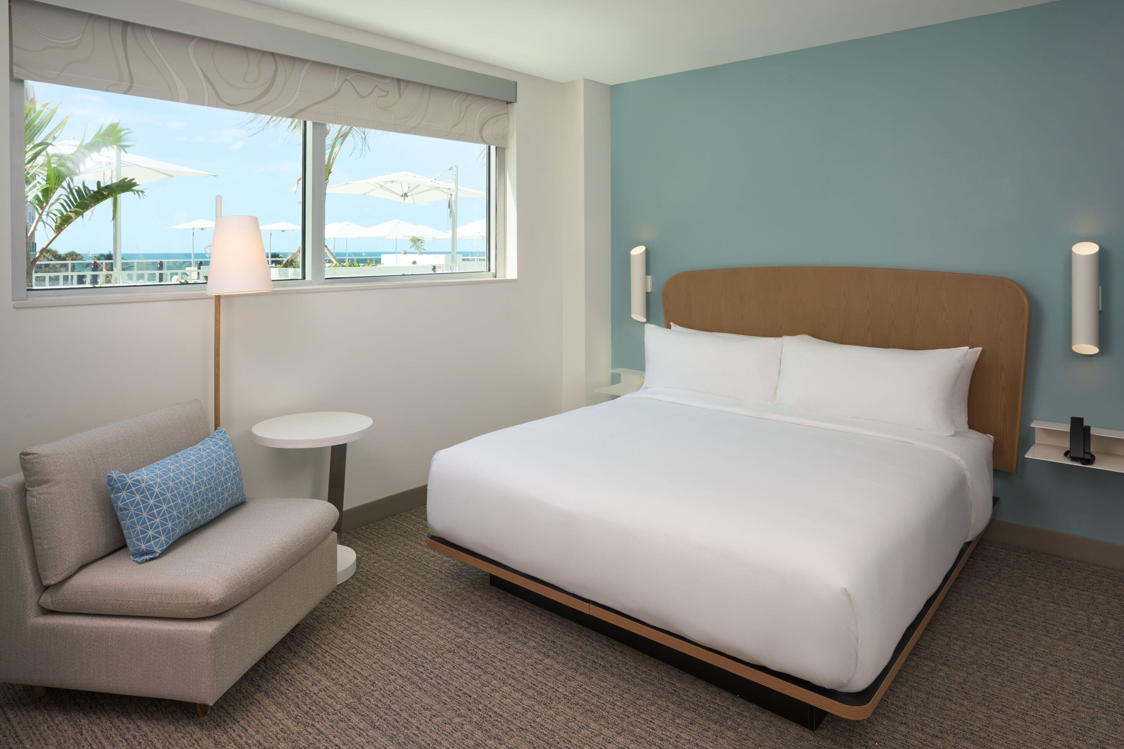 Habitación de hotel con cama, un pequeño sofá, una mesa redonda y vistas al océano a través de amplias ventanas.