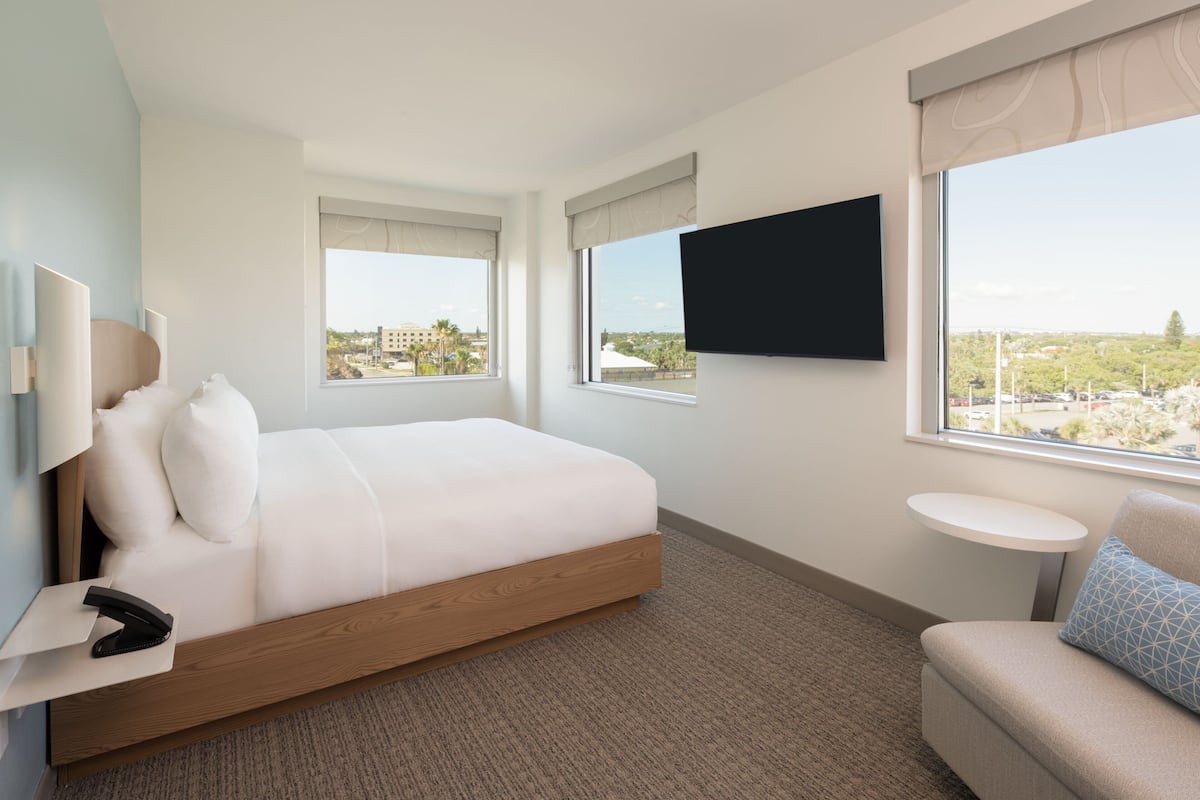 A hotel room with a bed, a TV mounted on the wall, a small table, and a window with a view of the city.
