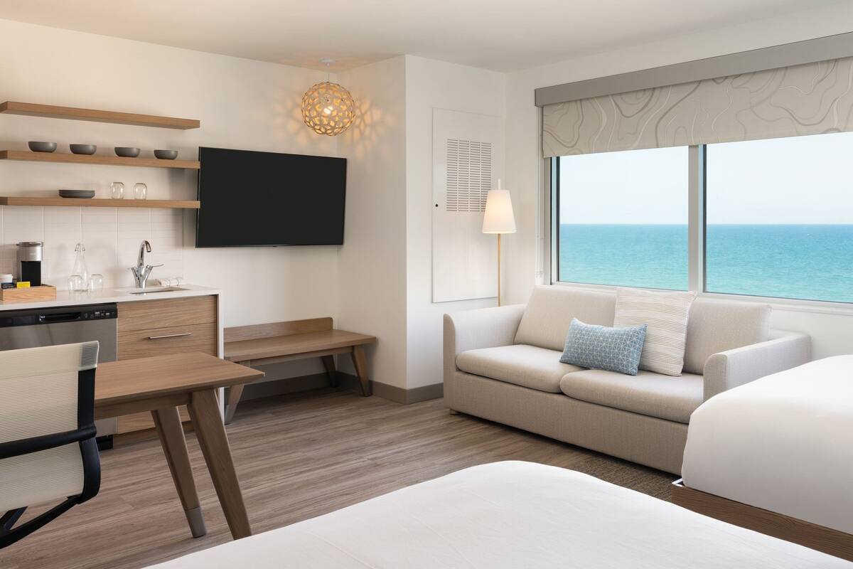 A modern hotel room with a kitchen area, a sofa, a bed, and a large window with a view of the sea.