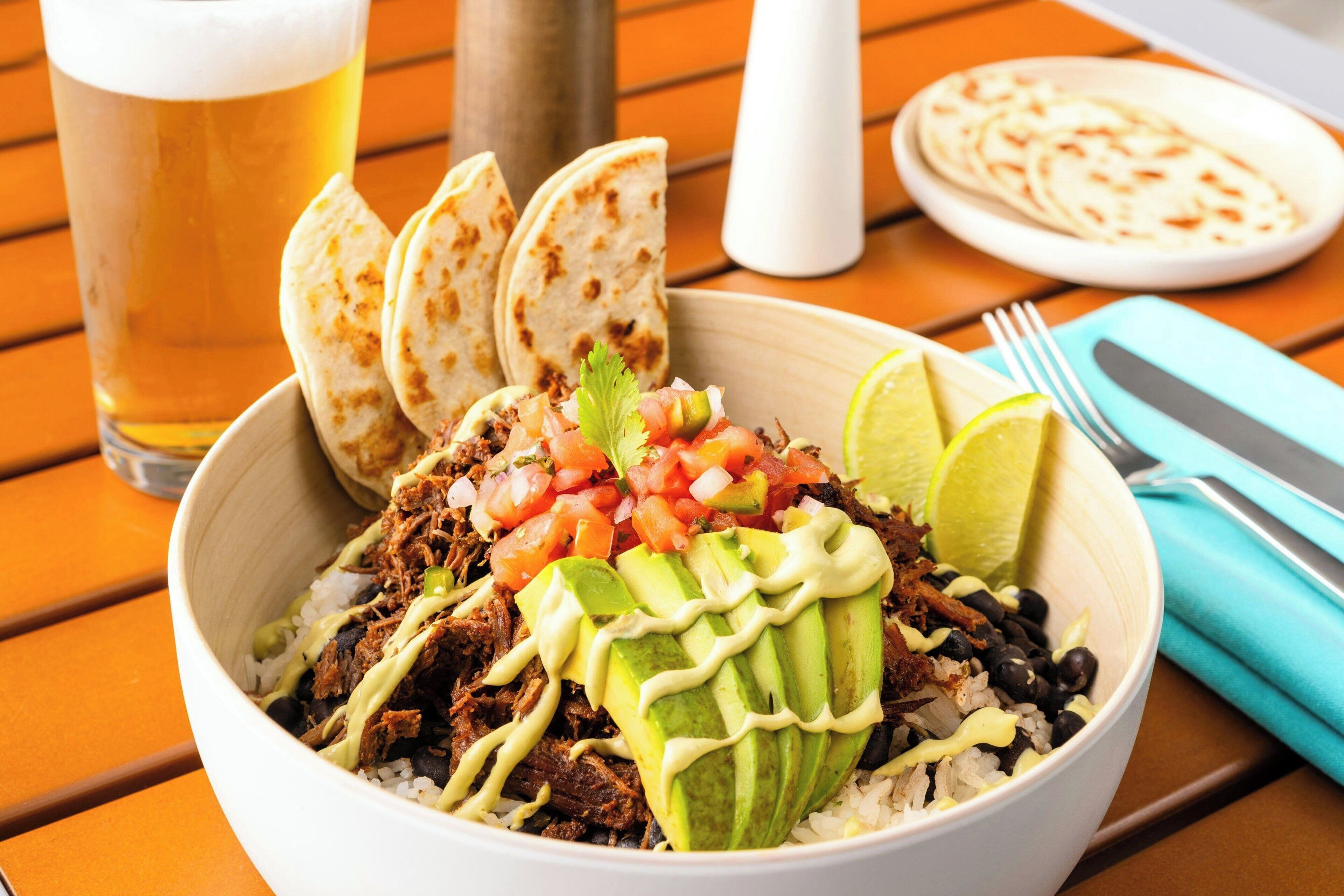 A bowl of shredded meat with avocado, black beans, and rice, garnished with lime and salsa, served with tortillas and a glass of beer on a wooden table.